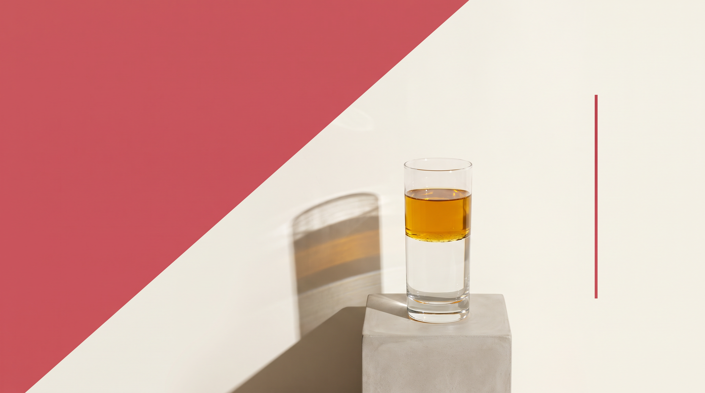 Un bicchiere di vetro con olio ambrato separato in due strati sopra l'acqua, su fondo diviso da una diagonale, metà rosso e metà bianco osso