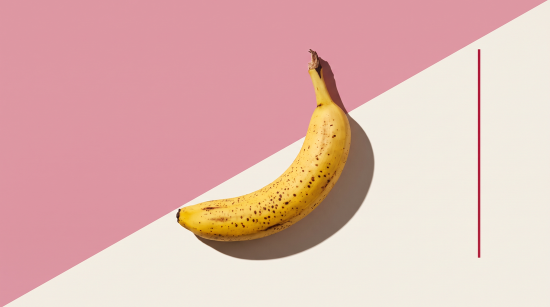Una banana matura con macchie scure sulla buccia gialla, su fondo diviso da una diagonale, metà rosa e metà bianco osso